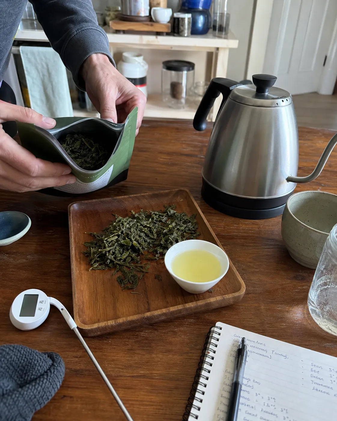 Hands checking green tea leaves beside a thermometer, kettle, cup, and resealable storage pouch